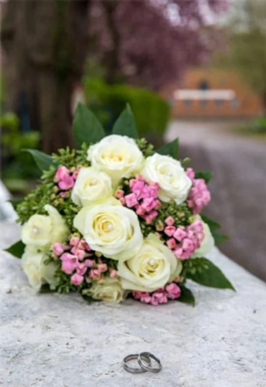 Brautstrauß aus weißen Rosen und rosa Bartnelken, davor zwei Eheringe